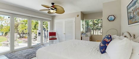A bedroom with a large bed, a ceiling fan, a view of the outdoors, and a door leading to a patio area.