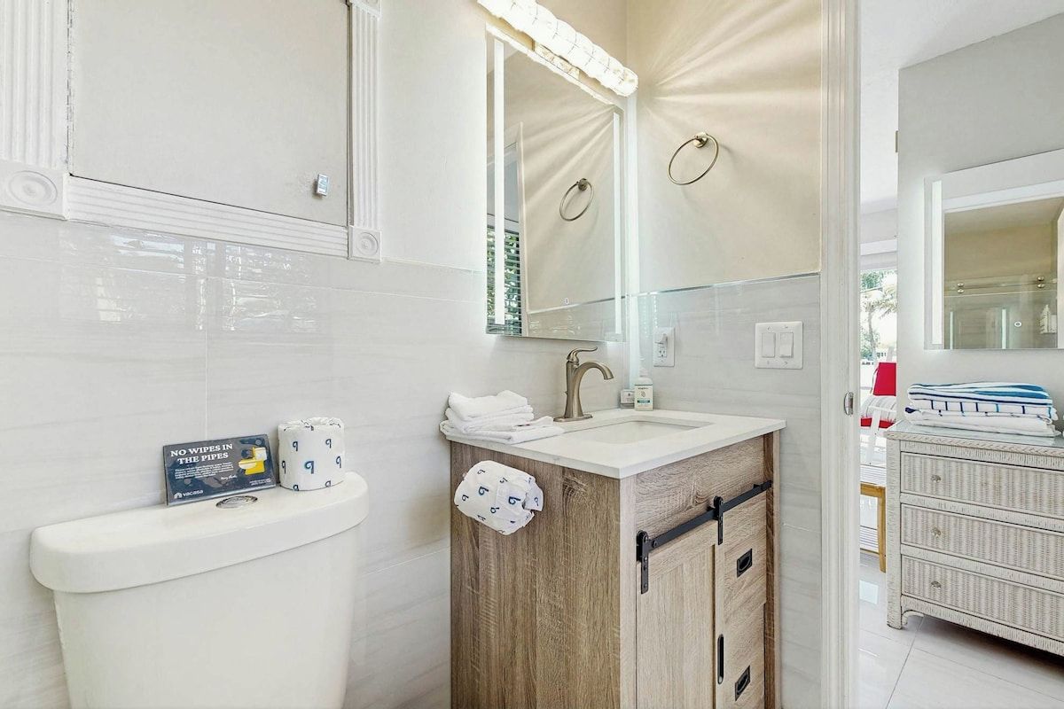 A bathroom with a toilet, sink, and mirror. There is a sign on the toilet, a towel rack, and a cabinet with drawers.