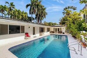 Una piscina con borde de azulejos, rodeada de palmeras y un edificio con grandes ventanales y un patio techado.