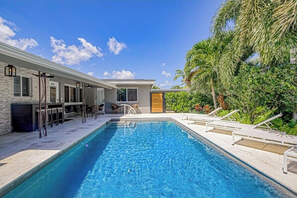 A swimming pool with lounge chairs, a bar area, and a door leading to a covered patio. - Designer Delight - Private Pool (Wilton Manors)