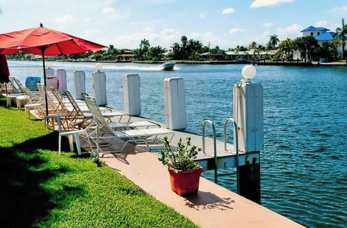 A waterfront pool area with lounge chairs and a red umbrella, a boat in the background, and a clear view of the water.