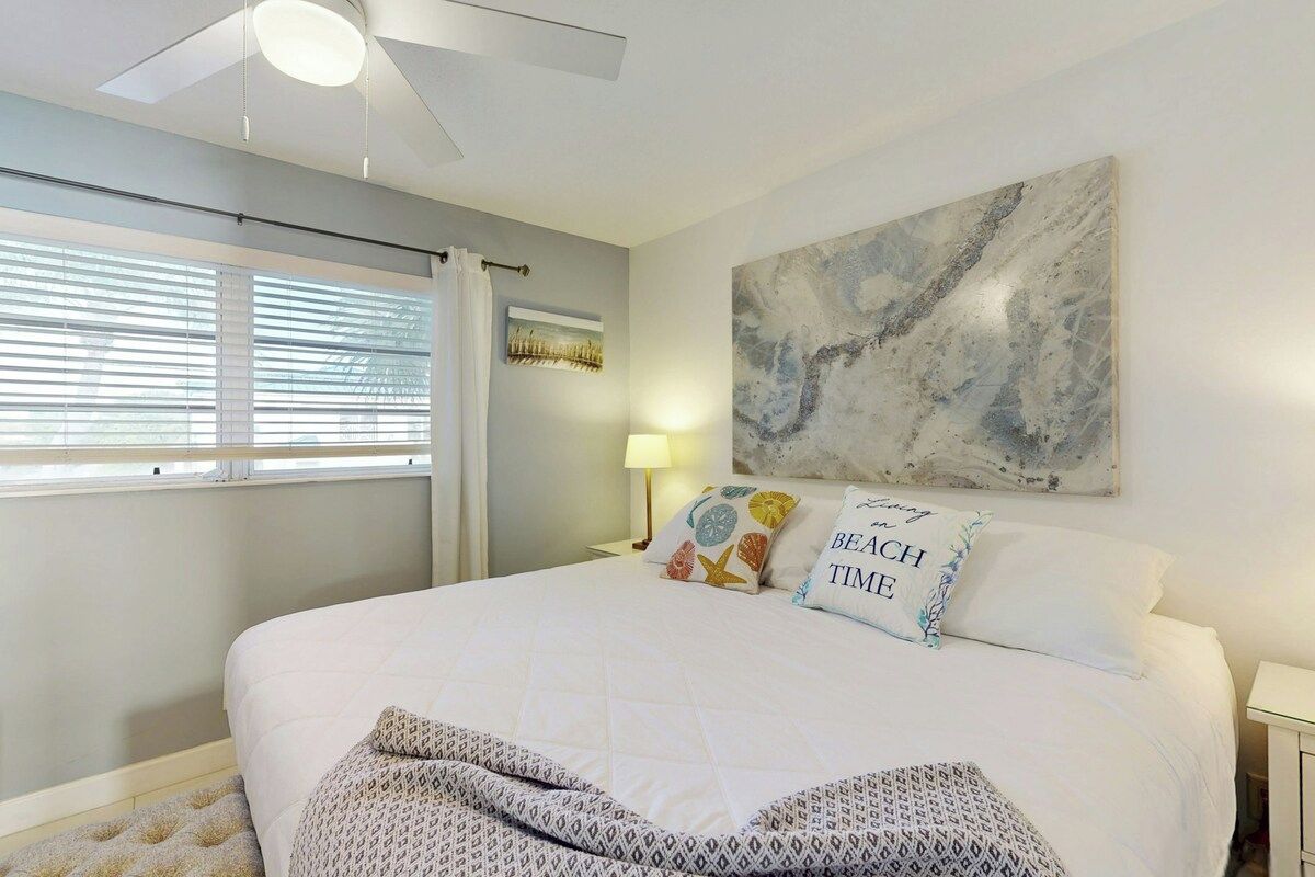 A bedroom with a large bed, a ceiling fan, a window with blinds, a wall-mounted artwork, and a bedside lamp.