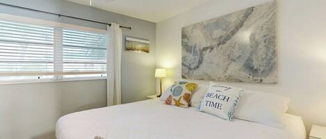 A bedroom with a large bed, a ceiling fan, a window with blinds, a wall-mounted artwork, and a bedside lamp.