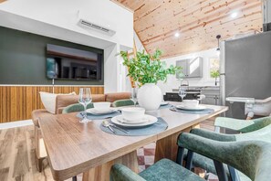 Un comedor moderno con una mesa de madera, sillas tapizadas en verde, un jarrón blanco grande con plantas verdes y un televisor de pantalla plana montado en la pared.
