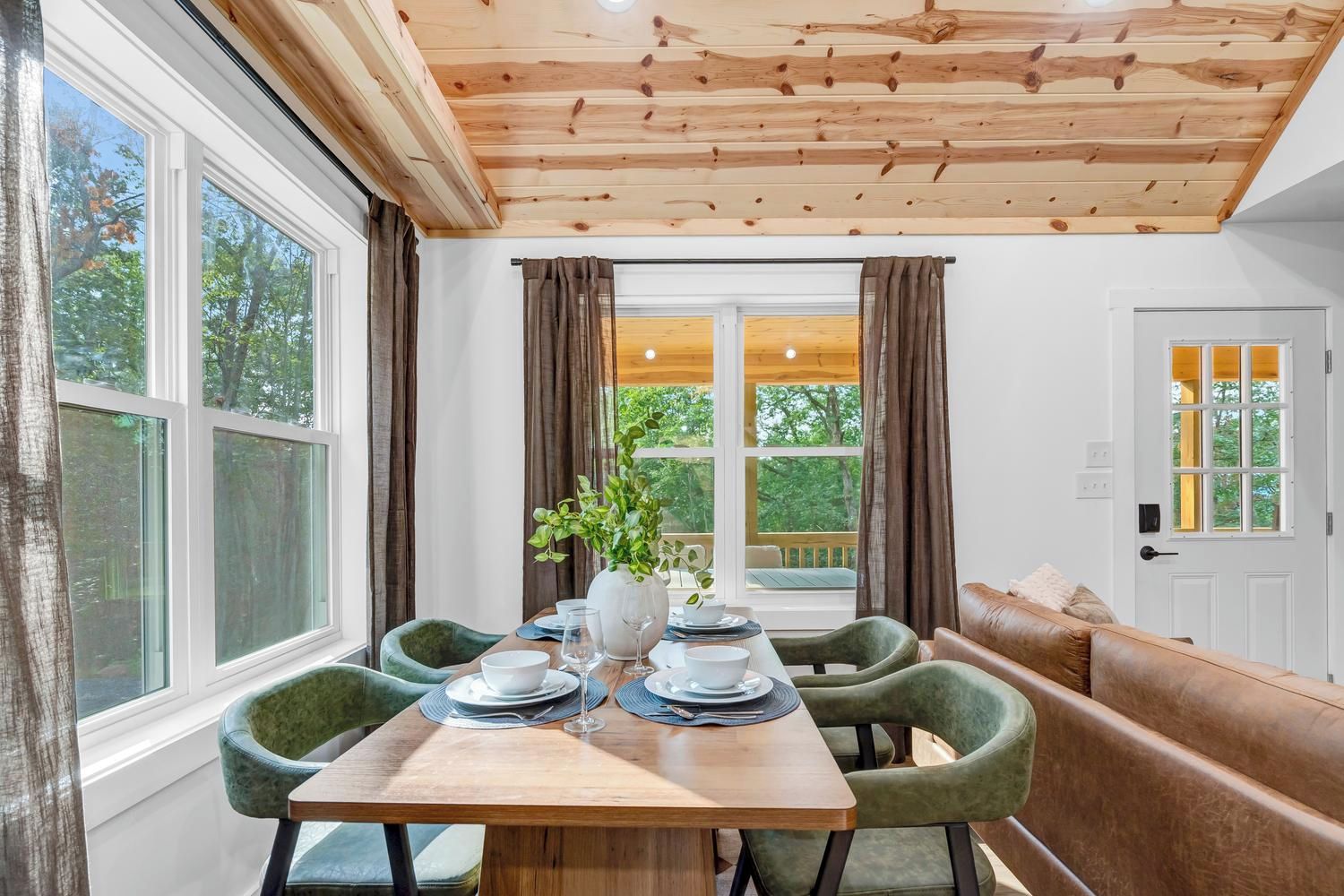 A dining area with a wooden table set for four, surrounded by green upholstered chairs, a bench, and a view of greenery outside.
