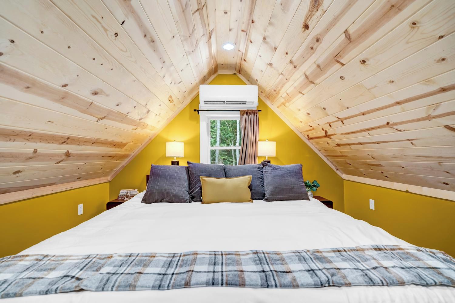 A room with a sloped ceiling, a bed with pillows, two bedside lamps, and a window with curtains.