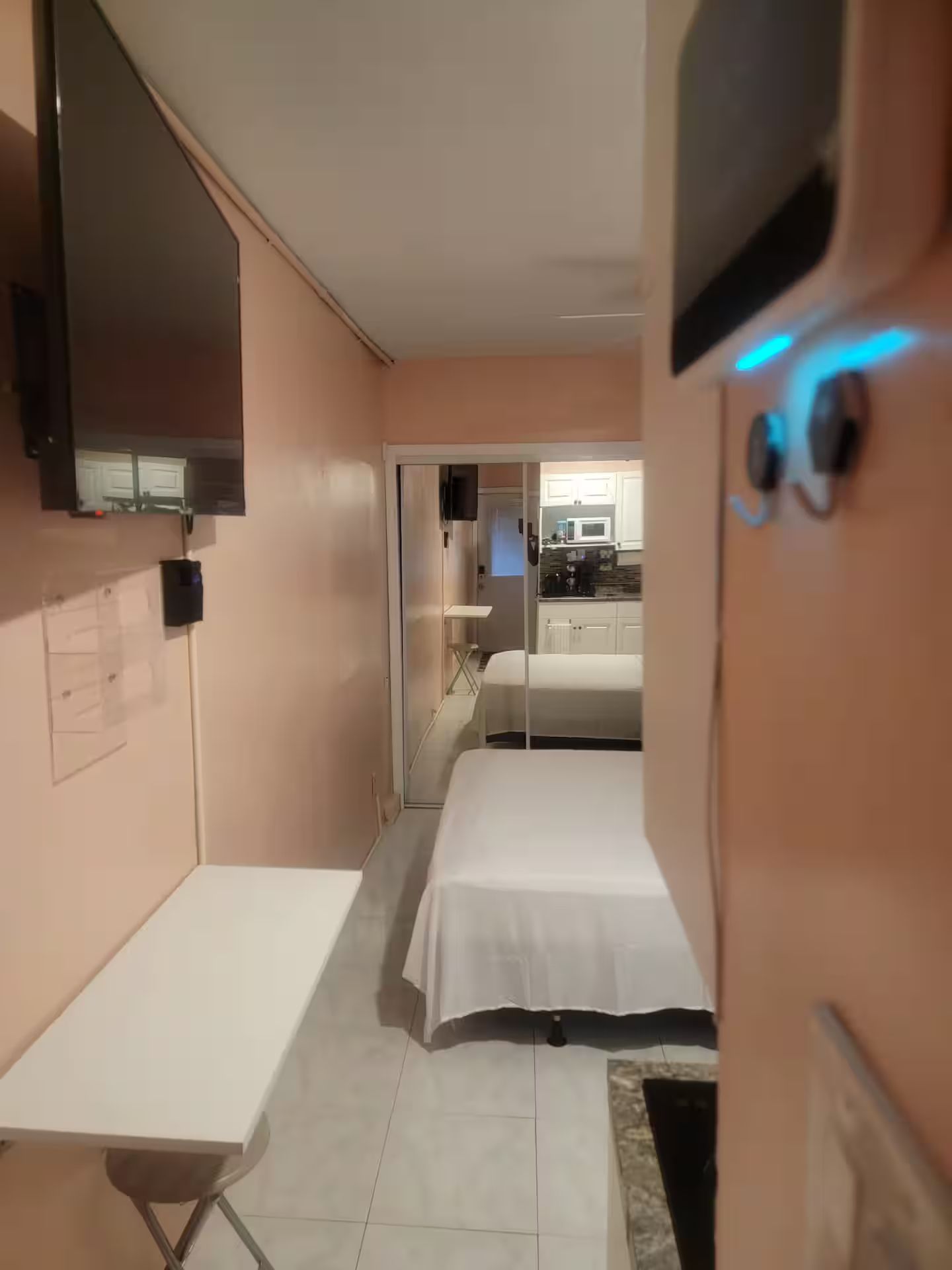 A hotel room with a bed, a desk, a television mounted on the wall, and a kitchenette in the background.