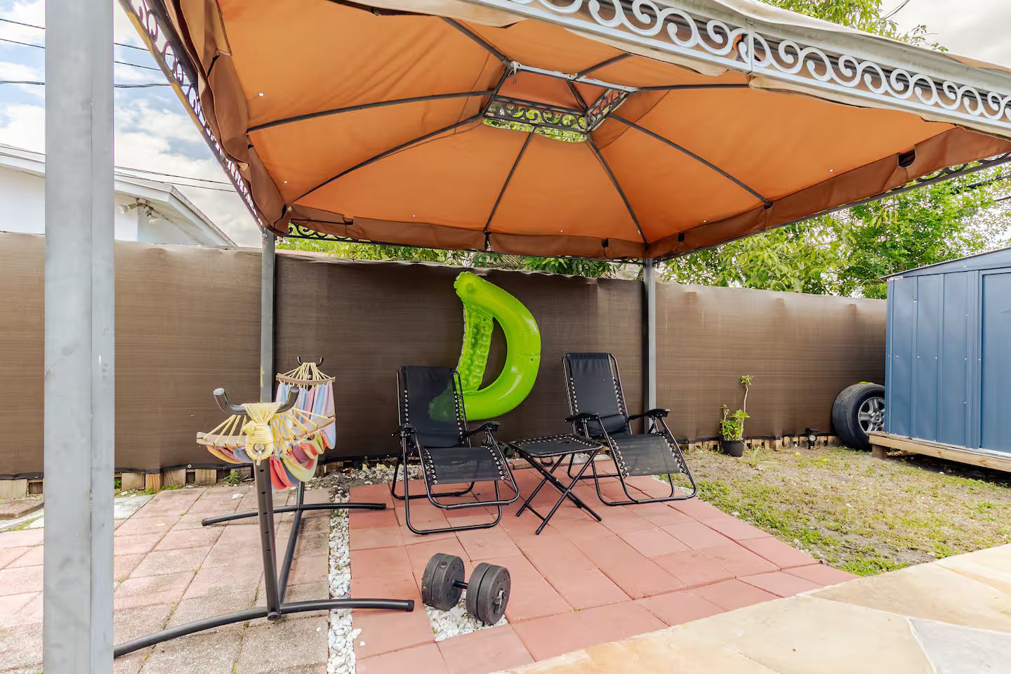 A patio with a large umbrella, two folding chairs, a green inflatable pool float, and a small table with a hanging rack.