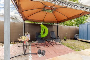 Une terrasse dotée d’un grand parasol, de deux chaises pliantes, d’un flotteur de piscine gonflable vert et d’une petite table avec un porte-objets suspendu.