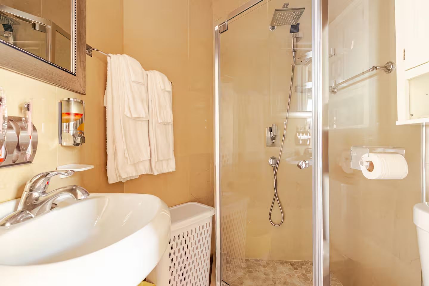 Un baño con lavamanos, espejo, toallero, ducha y dispensador de papel higiénico.