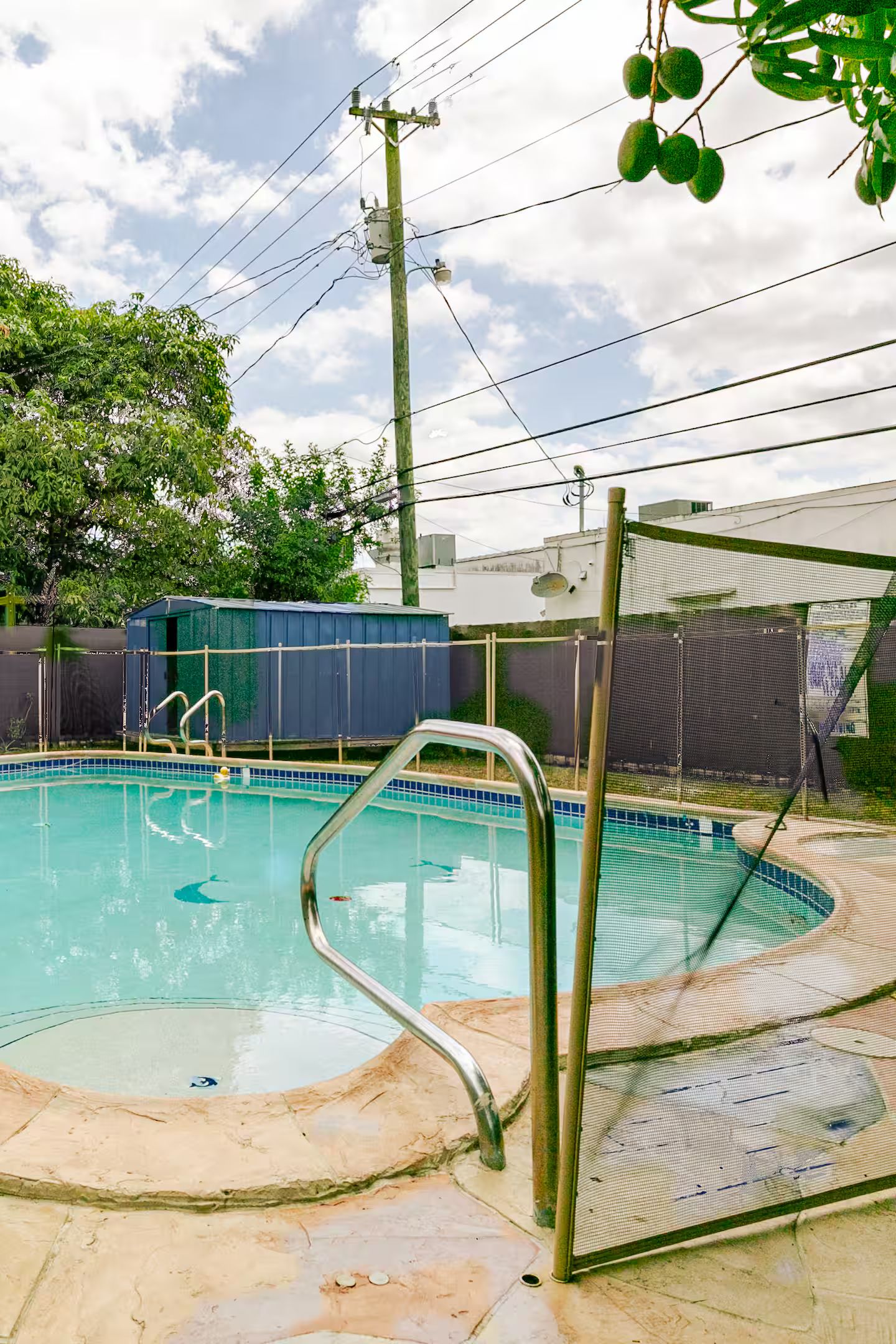 A swimming pool with a metal ladder and a covered area, surrounded by a fence and trees.