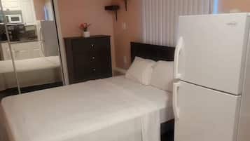 A bedroom with a bed, a white refrigerator, a dark dresser, and a window with blinds.