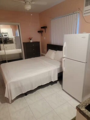 Une chambre à coucher avec un lit, un réfrigérateur blanc, une commode sombre et une fenêtre avec des stores.