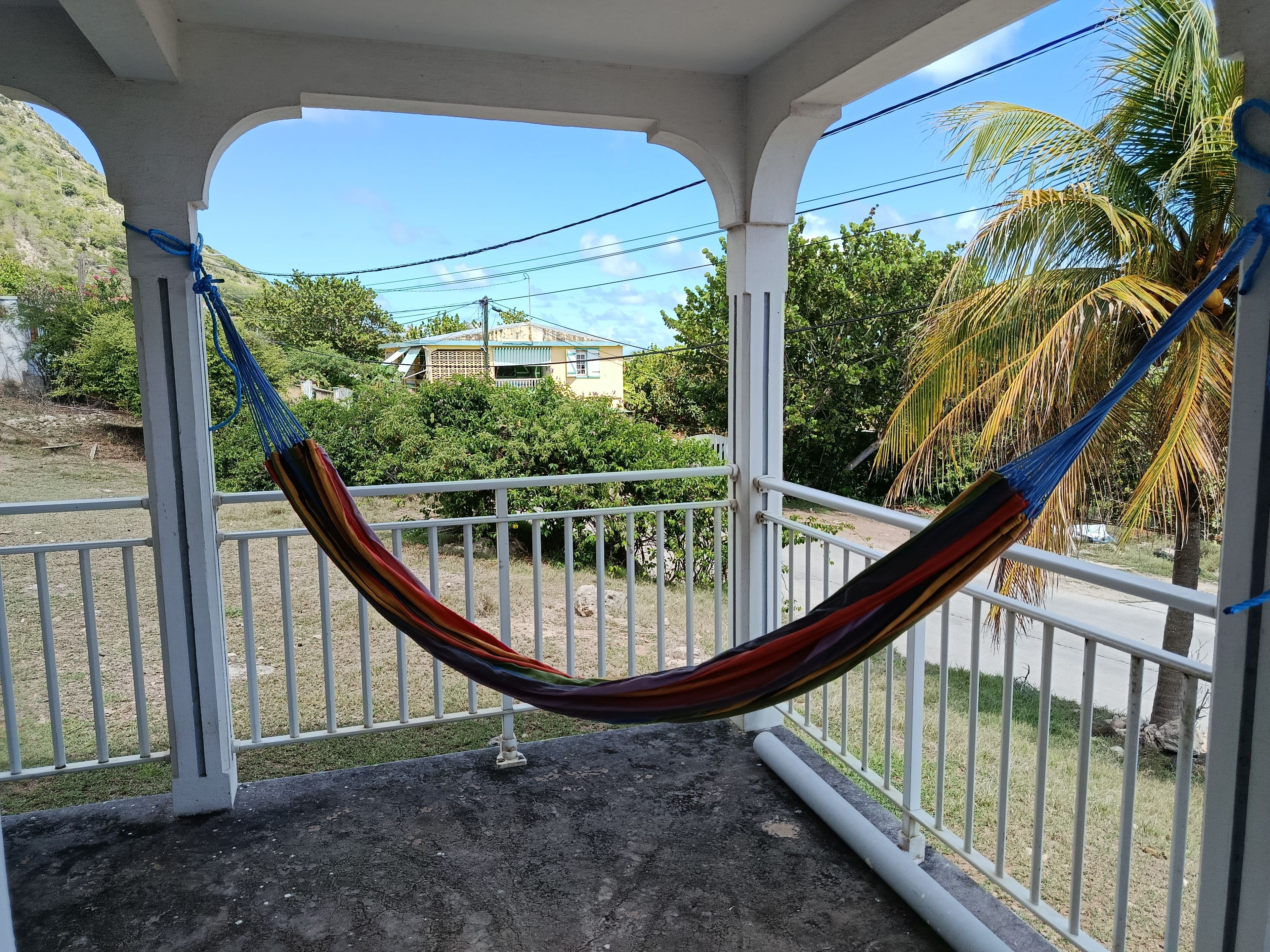 Un hamac est suspendu entre des balustrades blanches sur un balcon, d’où l’on peut admirer une maison et des palmiers.