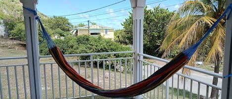 Una hamaca colgando entre barandas blancas en un balcón con vista a una casa y palmeras.