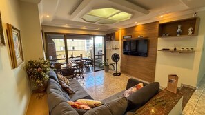 A living room with a sofa, a TV mounted on the wall, a fan, and a dining area with chairs and a table. - SHB - Churrasco - Piscina - 3 min praia (Guarujá)