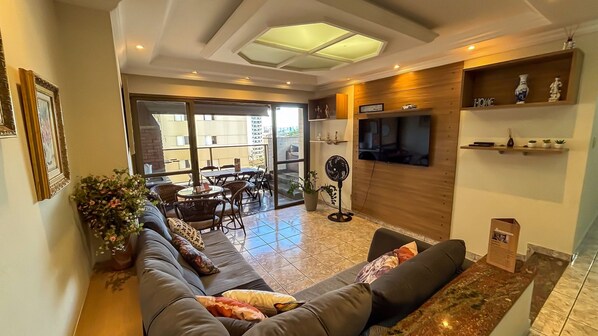 Una sala de estar con un sofá, un televisor montado en la pared, un ventilador y una zona de comedor con sillas y una mesa.