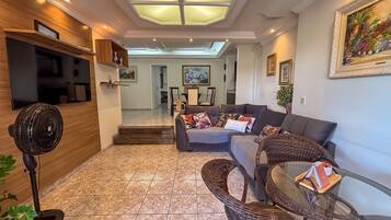 A living room with a sofa, a fan, a TV, and a dining area in the background.