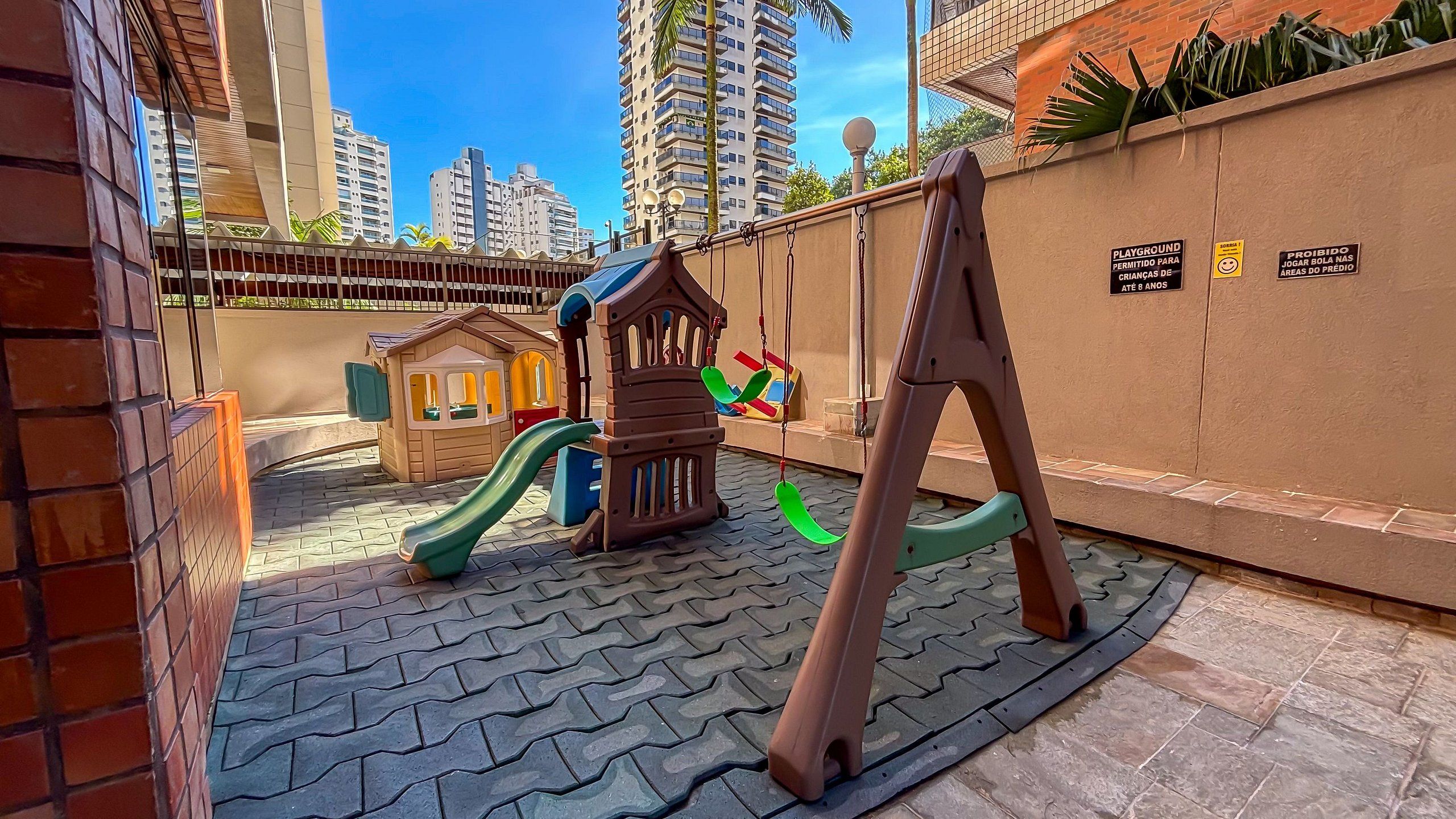 Un parque infantil con tobogán, columpios y casita, rodeado por un muro de ladrillo y una zona pavimentada.