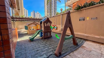 Un parque infantil con tobogán, columpios y casita, rodeado por un muro de ladrillo y una zona pavimentada.