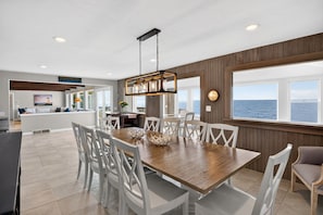 A spacious dining area with a wooden table and white chairs, a kitchen with a view, and a large window overlooking the sea. - Oceanfront Coastal Escape • Walk to Town • Private Beach (Harwich)