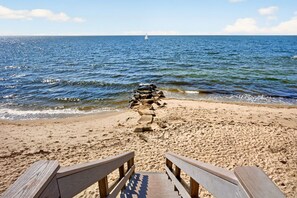 Une promenade en bois mène à une plage de sable, et on aperçoit un voilier au loin.