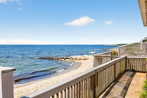 Eine Holzterrasse mit Blick auf einen Sandstrand, eine felsige Wellenbrecheranlage und einen klaren, blauen Himmel.