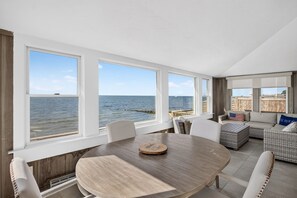 Une pièce dotée d’une grande fenêtre donnant sur la mer, d’une table ronde entourée de chaises et d’un coin salon avec un canapé et des coussins.