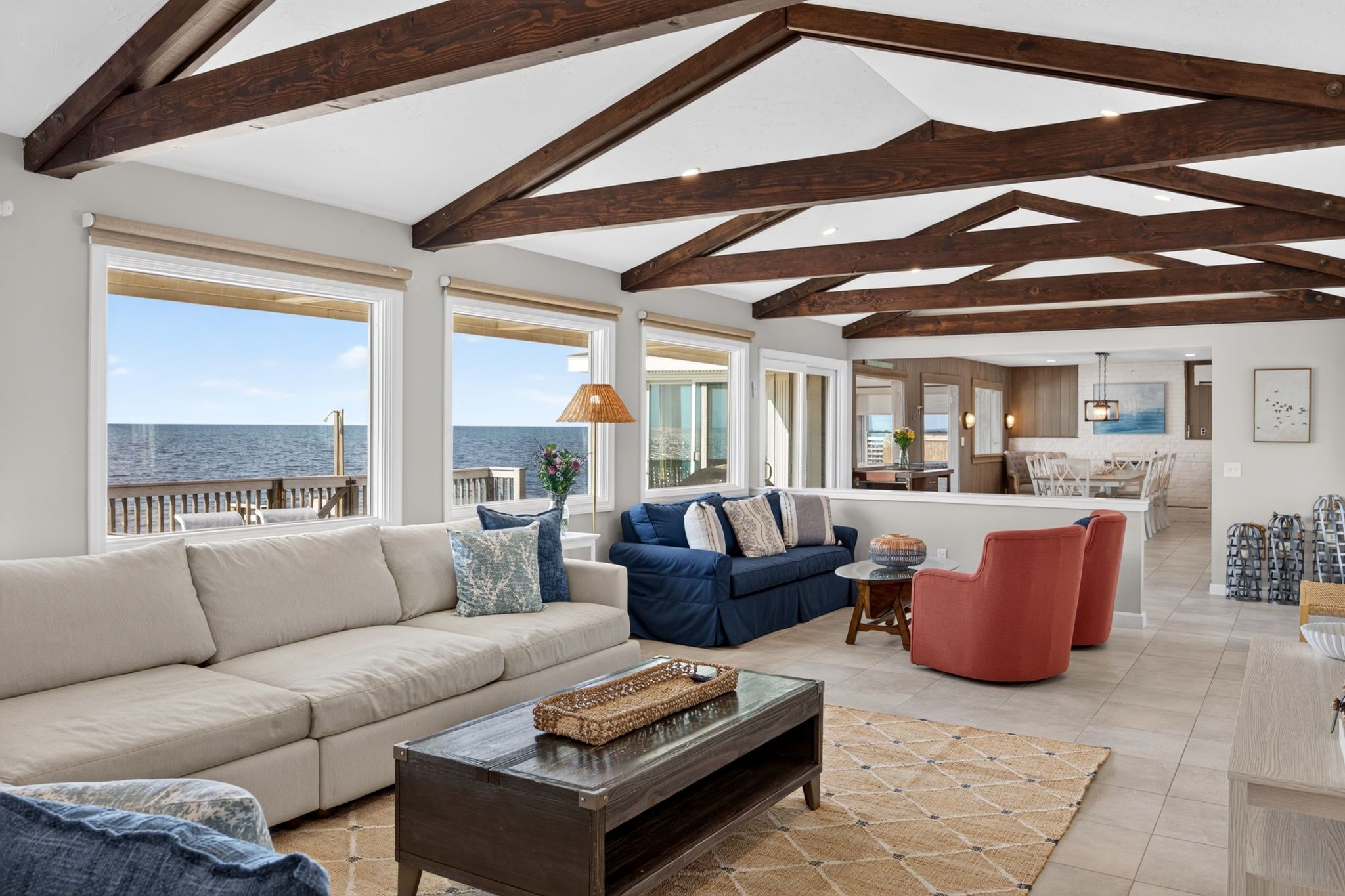 Un vaste espace de vie doté d’un plafond haut et de poutres apparentes, de grandes fenêtres offrant une vue sur la mer, un coin salon avec un canapé et des fauteuils, et un coin repas avec une table et des chaises.