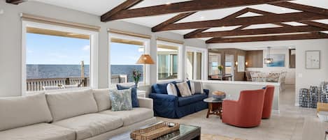 Un vaste espace de vie doté d’un plafond haut et de poutres apparentes, de grandes fenêtres offrant une vue sur la mer, un coin salon avec un canapé et des fauteuils, et un coin repas avec une table et des chaises.