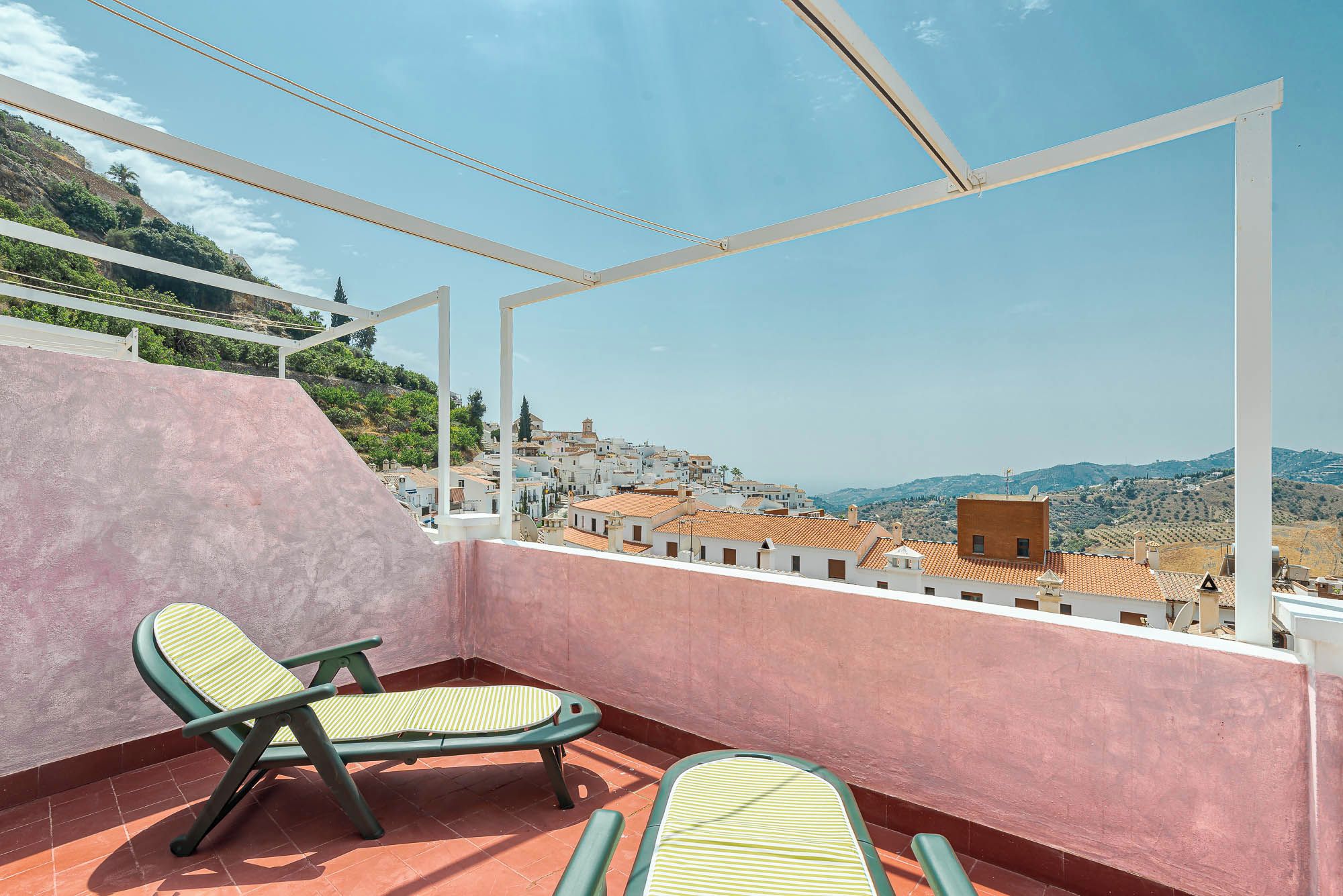 A balcony with two lounge chairs, a view of a town with white buildings, and a hilly landscape in the distance.