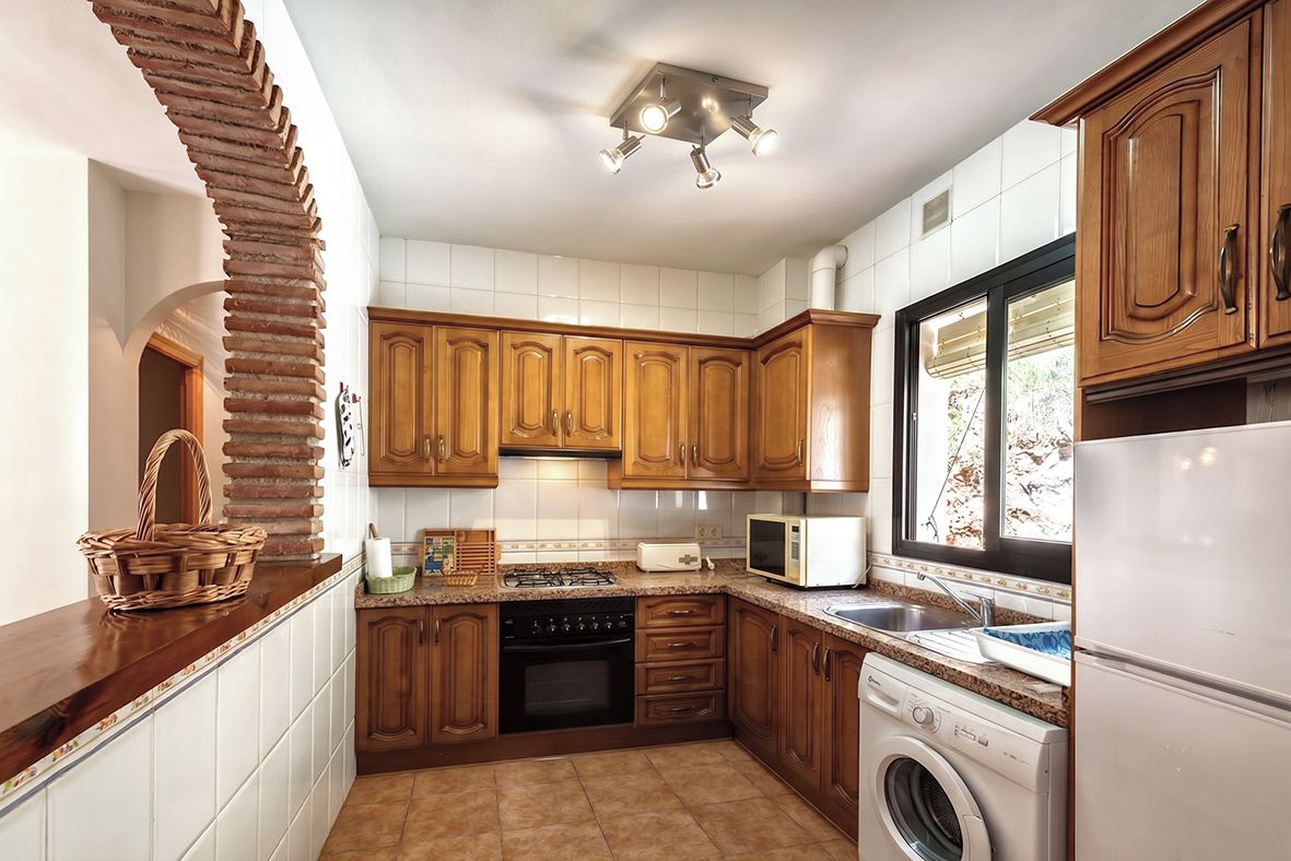 A kitchen with wooden cabinets, a microwave, a washing machine, and a window with blinds.