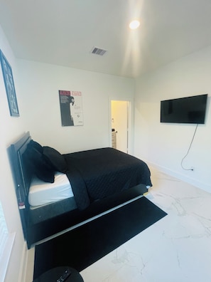 Quarto com cama, televisão de tela plana na parede, pôster na parede e porta que dá para outro cômodo.