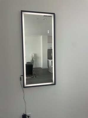 A mirror with a black frame mounted on a wall, reflecting a room with a dark wooden floor, a white wall, a small table with a vase, and a kitchen area with cabinets and a refrigerator. - Luxury Living Collection 
Black & White Edition (Houston)
