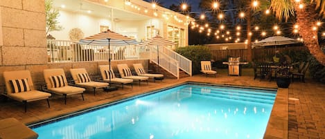 A swimming pool with lounge chairs and umbrellas, surrounded by a stone wall and string lights.