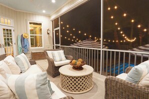 A covered outdoor seating area with wicker furniture, a round table with decorative items, and string lights. - East Beach Estate Between Ocean & Estuary | Salt Pool + Spa | Shuffleboard Table (St. Simons Island)