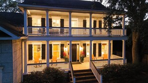 A two-story building with a balcony, a staircase leading to the entrance, and a well-lit porch area. - East Beach Estate Between Ocean & Estuary | Salt Pool + Spa | Shuffleboard Table (St. Simons Island)