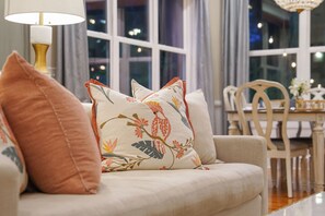 Een bank met bloemenkussens en een tafel met stoelen in een kamer met grote ramen.