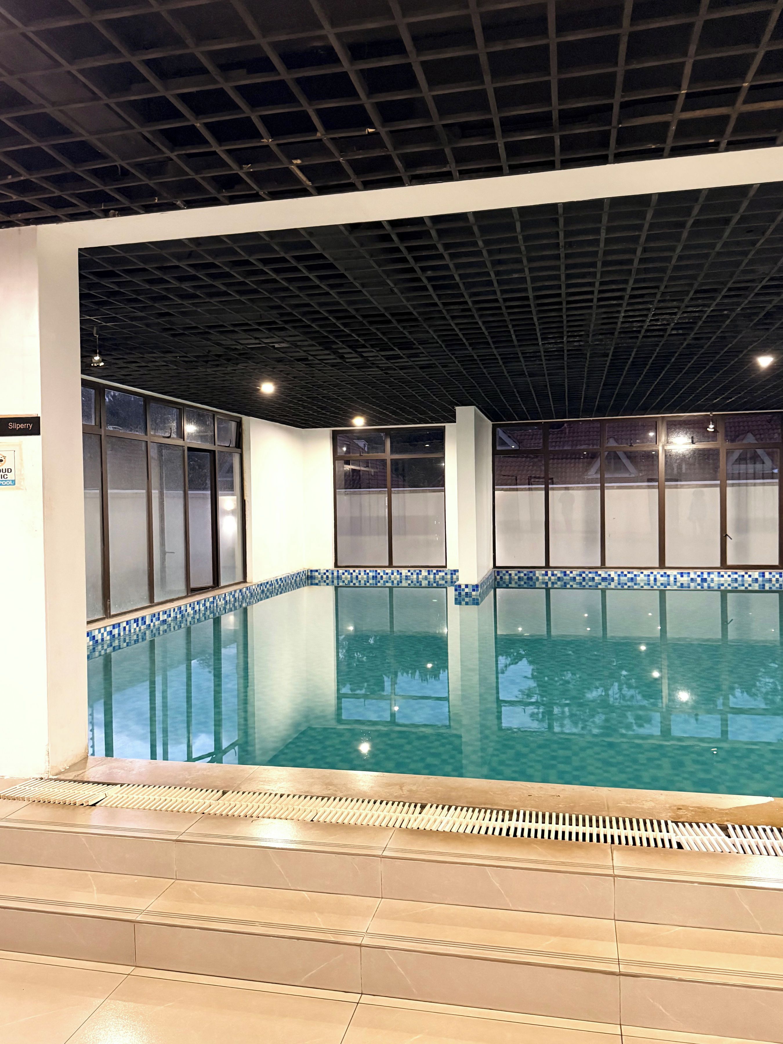 An indoor swimming pool with clear water, tiled steps leading into the pool, and a ceiling with a grid pattern.