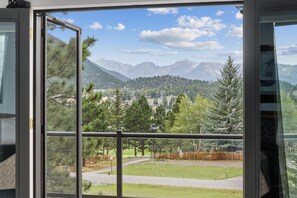 A view from a window with a metal railing, overlooking a mountainous landscape with trees and a road. - Charming Estes Lake Haus with Stunning Mountain Views & Walk to Lake Estes (Estes Park)