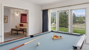 A pool table with balls and cues, a sofa, a red chair, and a view of a green landscape through large windows. - Charming Estes Lake Haus with Stunning Mountain Views & Walk to Lake Estes (Estes Park)