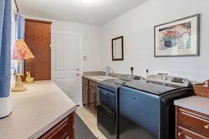 A compact kitchen with a sink, stove, and refrigerator. There's a door with a wooden panel, a small table with a lamp, and a framed artwork on the wall. - Charming Estes Lake Haus with Stunning Mountain Views & Walk to Lake Estes (Estes Park)