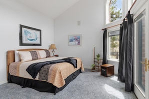 Ein Schlafzimmer mit einem Bett, zwei gerahmten Kunstwerken, einer Nachttischlampe, einem kleinen Tisch mit einem Lautsprecher und einem Fenster mit Vorhängen.