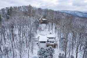 Une maison moderne à deux étages, entourée d’une forêt où les arbres sont couverts de neige.
