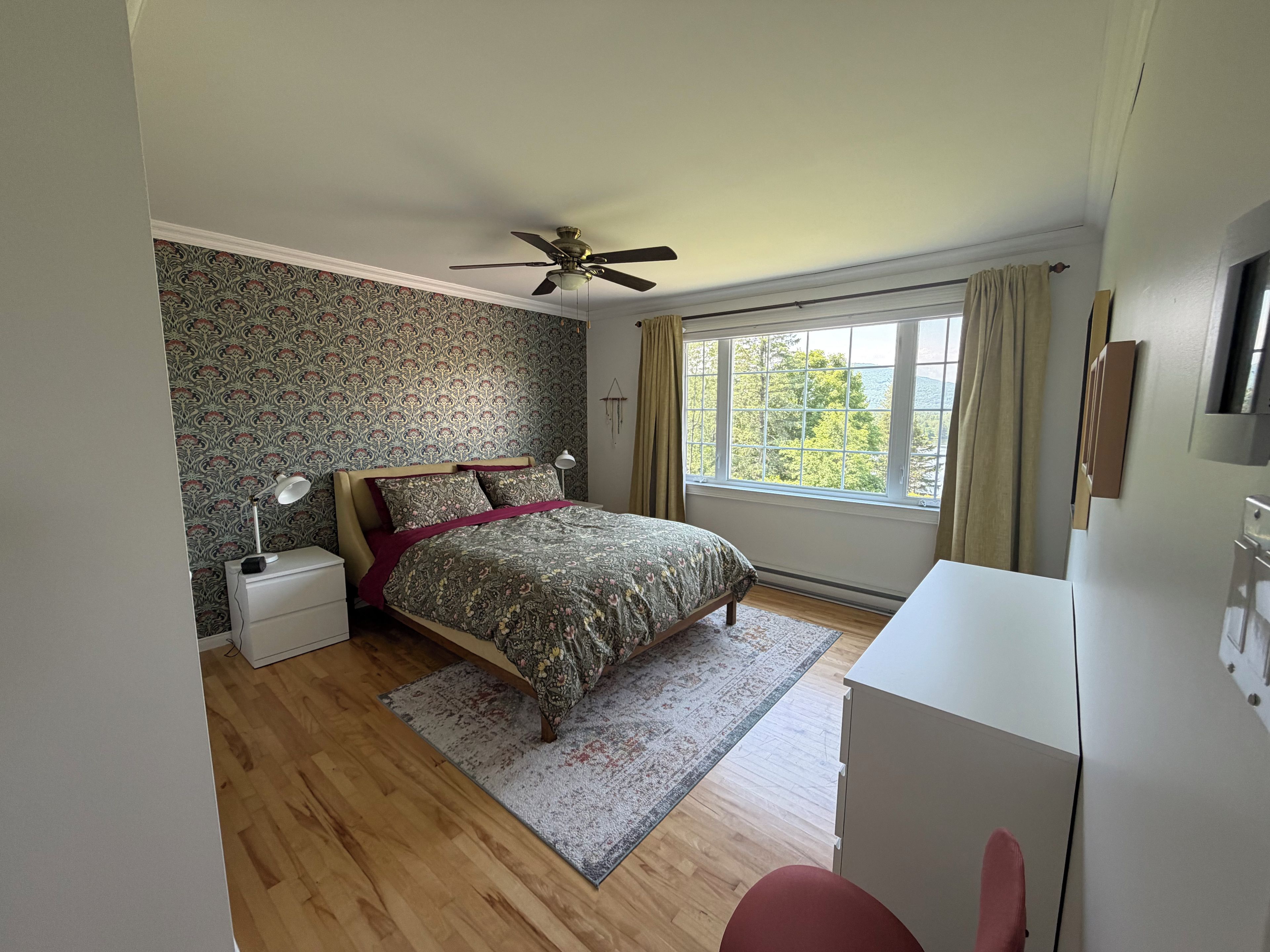 A bedroom with a bed, a desk, a ceiling fan, a window with curtains, and a patterned wallpaper.