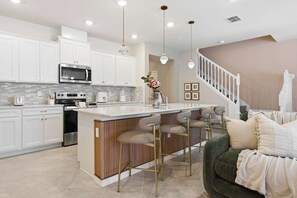 A modern kitchen with white cabinets, stainless steel appliances, a central island with bar stools, and a staircase leading to a living area. - 16217 Toy Story & Mickey Rooms! Windsor Cay 5BR (Clermont)