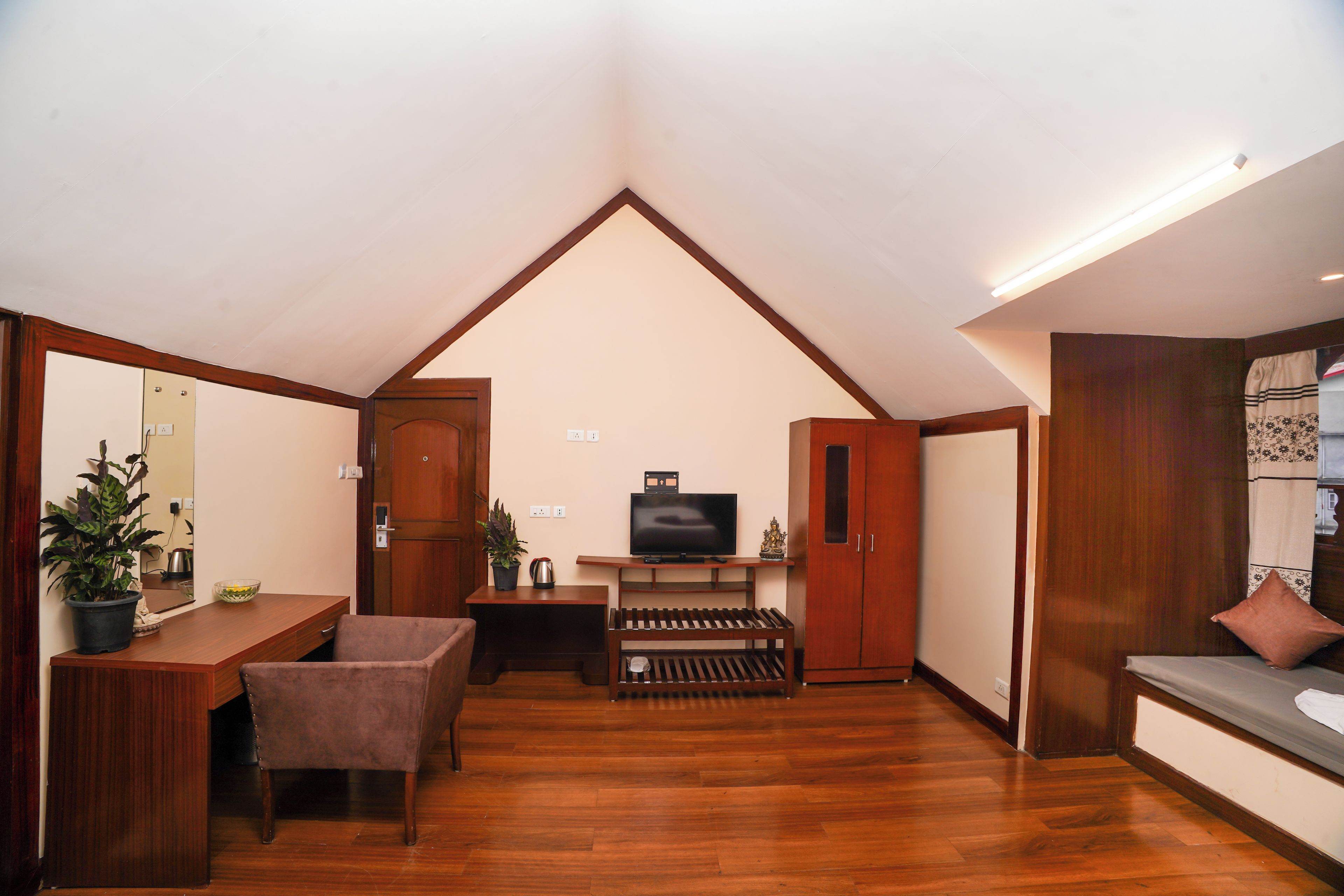 A room with a sloped ceiling, wooden flooring, a television, a sofa, a table with chairs, and a potted plant.