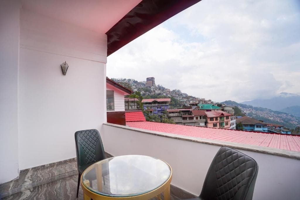 A balcony with a glass table and two chairs, overlooking a residential area with colorful houses and a distant hill.