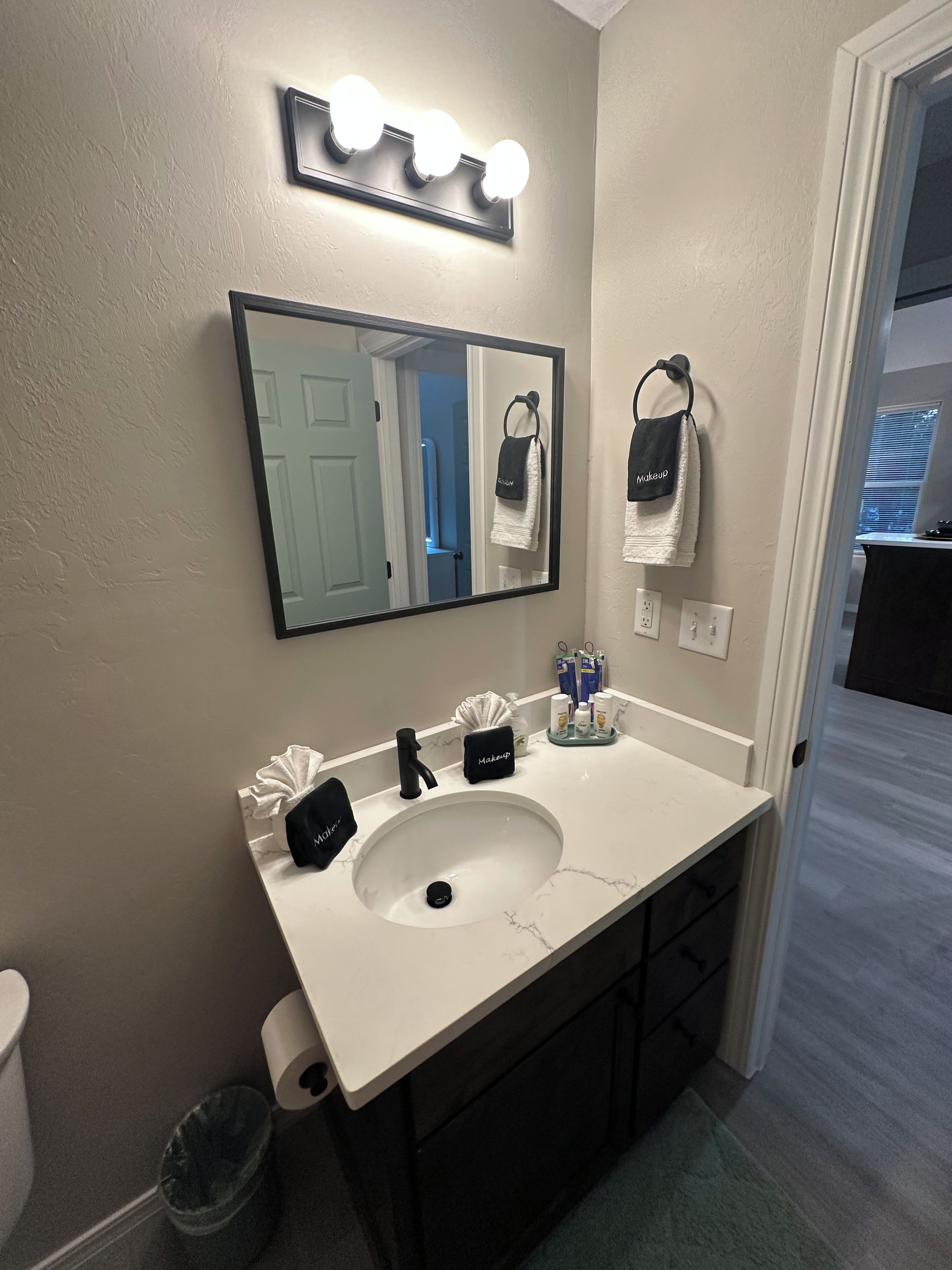 A bathroom with a white sink, a mirror with lights, a towel rack, and a trash can.