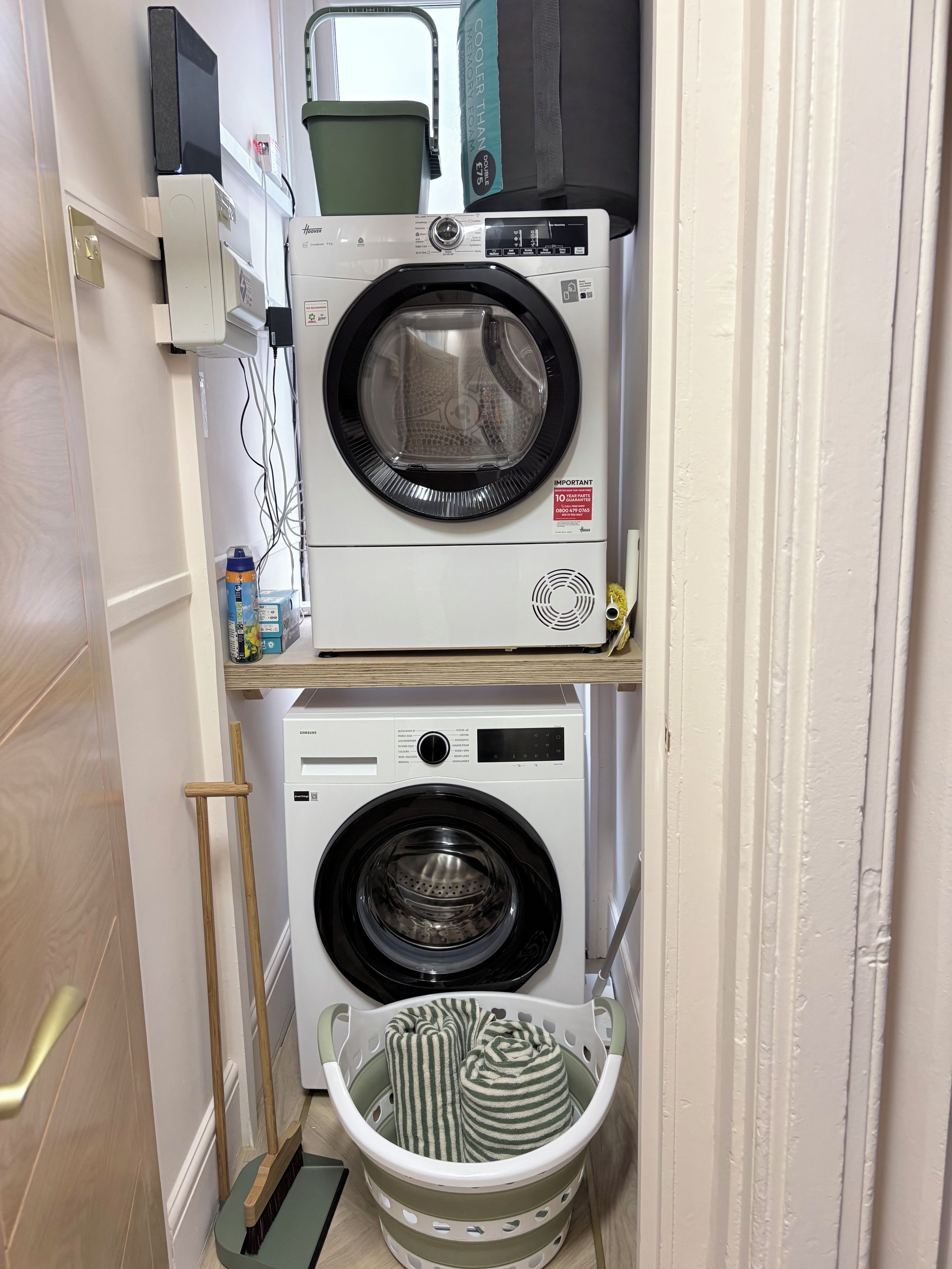 A compact laundry area with two stacked washing machines, a drying rack, and a broom.